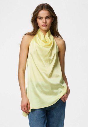 Jeune femme portant un haut sans manches en satin jaune pâle de type halter et un jean bleu, debout devant un fond blanc uni.