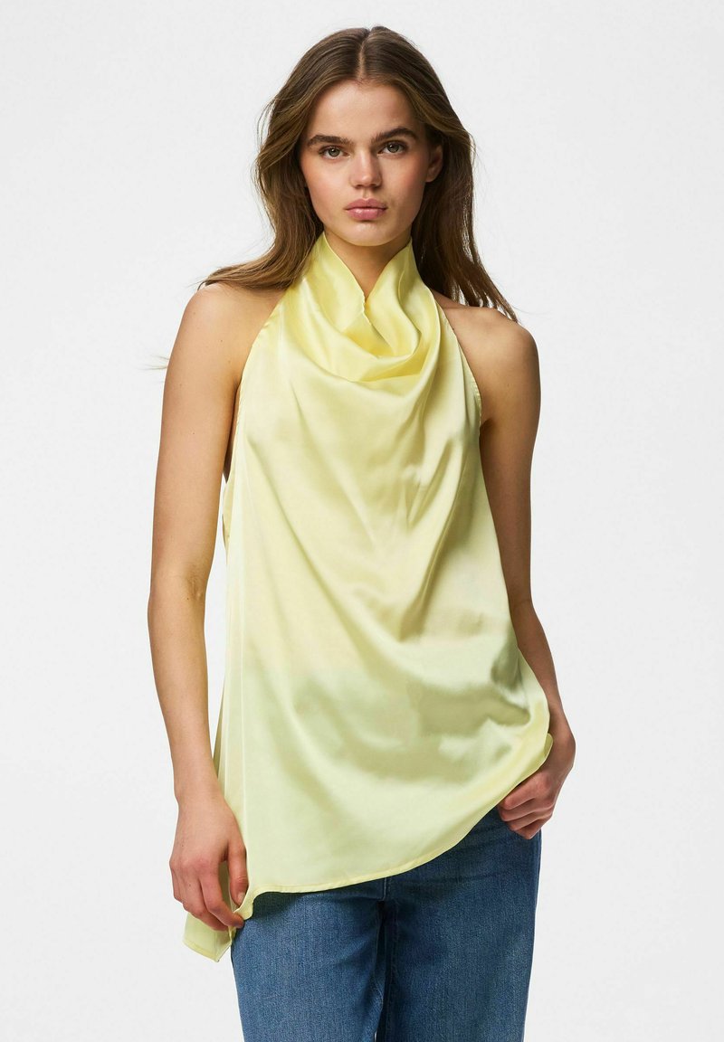Jeune femme portant un haut sans manches en satin jaune pâle de type halter et un jean bleu, debout devant un fond blanc uni.