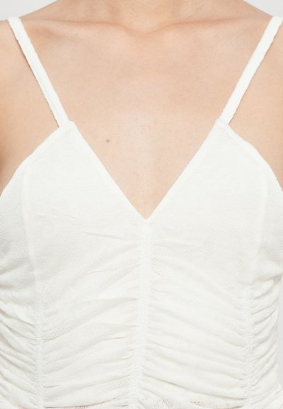 Haut sans manches blanc avec un décolleté en V, présentant un tissu texturé froncé et des bretelles fines. Le design met en valeur une silhouette lisse et ajustée.