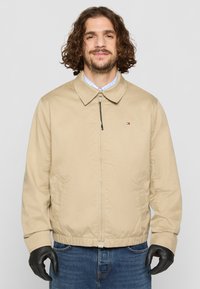 Bež bomber jakna z ovratnim izrezom, sprednjim zadrgo, stranskimi žepi in elastičnimi manšeti. Nosi se preko svetlo modre karirane srajce.