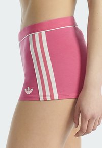 Shorts Adidas roses avec trois bandes blanches et le logo trèfle sur le côté gauche, portés par une personne montrée de la taille aux pieds.