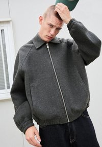 Giacca grigia con zip, colletto, polsini e vita elastici. Realizzata in tessuto testurizzato, esposta su uno sfondo neutro.