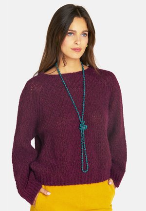 Frau mit langen braunen Haaren, die einen strukturierten violetten Pullover, eine türkisfarbene Perlenkette und senfgelbe Hose trägt, Hände in den Taschen, schaut nach links.