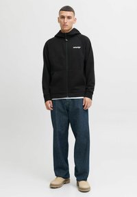 Zwarte zip-up hoodie met een voorzak en raglanmouwen, met een logo op de borst. Gedragen met blauwe baggy jeans en beige schoenen.