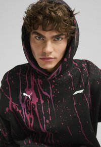 Sudadera negra con patrón de salpicaduras rosas, que cuenta con un logo en la parte frontal y capucha. Hecha de tela suave con un corte relajado.
