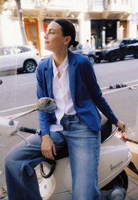 Asmuo mėlynu švarku ir balta palaidine sėdi ant balto motorolerio. Matosi šviesiai skalbti džinsai ir sklandžios, elegantiškos motorolerio formos.