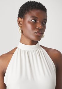 Haut blanc à col halter avec des plis. Le mannequin porte des boucles d'oreilles en or et perles, avec un design texturé. Tissu lisse, silhouette ajustée.