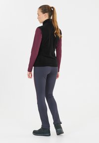 Sort fleece vest over en bordeaux langærmet bluse, parret med mørkegrå slim-fit bukser og sorte vandrestøvler. Ensfarvet baggrund.