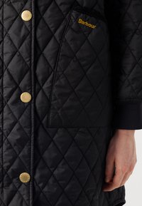 Zwarte gewatteerde jas met ruitpatronen, gouden drukknoopsluitingen en een zijzak met "Barbour" geborduurd in gouden draad.