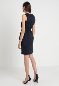 Granatowa sukienka bez rękawów z dopasowaną talią i detalem w postaci paska, gładka tekstura, do kolan, połączona z czarnymi butami na obcasie.