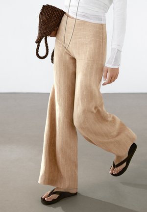 Pantaloni in lino beige a gamba larga con righe verticali, abbinati a ciabatte nere e una borsa in tessuto marrone. Design semplice e rilassato.