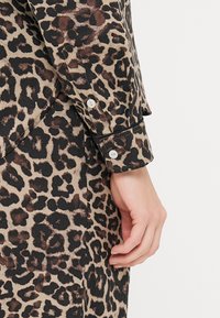 Leopardmønstret skjorte med lange ærmer, der har en knaplukning ved håndleddet. Materialet har en glat tekstur med nuancer af tan, sort og brun.