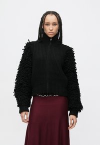 Černý svetr se zipem s žebrovaným tělem a texturovanými, nadýchanými rukávy, nošený přes vínovou sukní s volánkovým lemem.