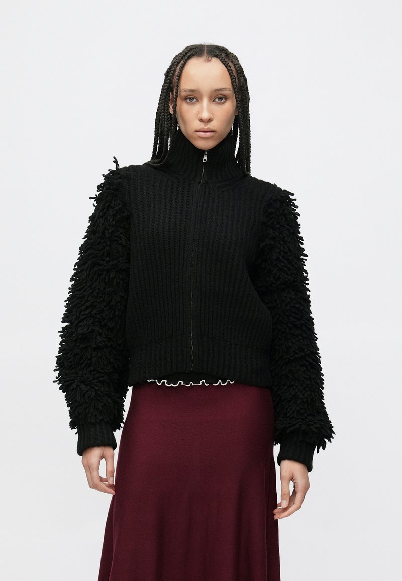 Černý svetr se zipem s žebrovaným tělem a texturovanými, nadýchanými rukávy, nošený přes vínovou sukní s volánkovým lemem.
