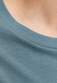 Teal crew-neck t-shirt med struktureret strik. Stoffet er glat, og kraven har et ribbet design. Nærbillede af halsudskæringen.