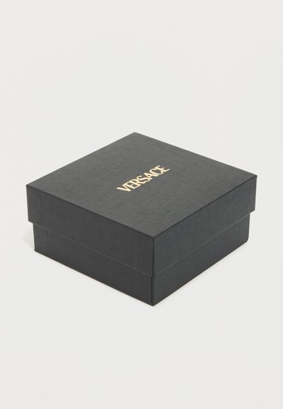 Μαύρο τετράγωνο κουτί με υφή, με χρυσό λογότυπο "Versace" τοποθετημένο στο κέντρο του καπακιού, επάνω σε απλό λευκό φόντο.