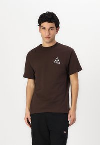 Camiseta de algodón marrón con escote redondo y mangas cortas, que presenta un logo geométrico triangular blanco en la parte izquierda del pecho.