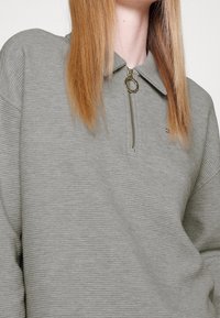 Sudadera gris claro con cremallera y cuello, con textura acanalada, herrajes dorados en la cremallera y un emblema en el lado izquierdo del pecho.