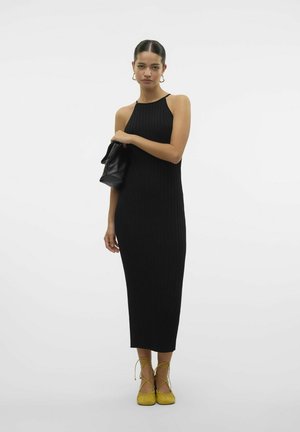 Robe midi noire côtelée avec une encolure haute. Le mannequin tient un sac noir texturé. Des ballerines jaunes vives à lacets avec une finition douce complètent le look.