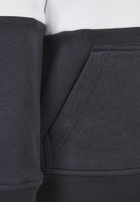 Felpa blu navy scuro con una striscia orizzontale bianca, texture morbida, polsini a coste e una tasca frontale sul lato inferiore destro.