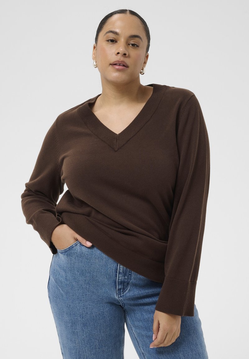 Femme avec un pull marron en V et un jean bleu, posant avec une main dans la poche devant un fond uni.