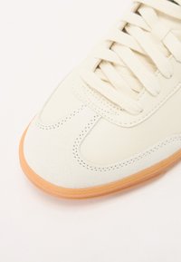 Krémszínű bőr felsőrésszel és velúr részletekkel rendelkező sneaker, kerek orral, lapos fűzős megoldással és világosbarna gumitalppal.