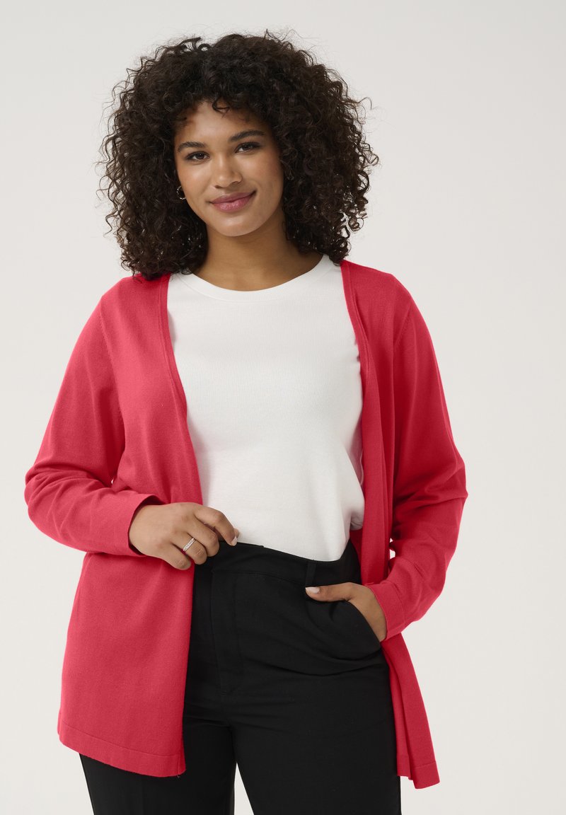 Cardigan en tricot rouge avec des manches longues, design à l'avant ouvert. Porté sur une chemise blanche et associé à un pantalon noir. Texture lisse, coupe décontractée.
