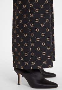 Black high-heeled shoes with a pointed toe and slim heel, paired with patterned trousers featuring a diamond and circle design in tan.