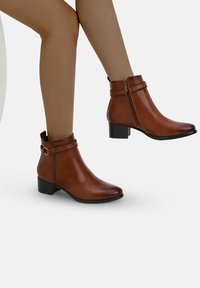 Botas de cuero marrón hasta el tobillo con punta redondeada, tacón cuadrado y correa decorativa con hebilla. Cuenta con una cremallera lateral para facilitar su uso.