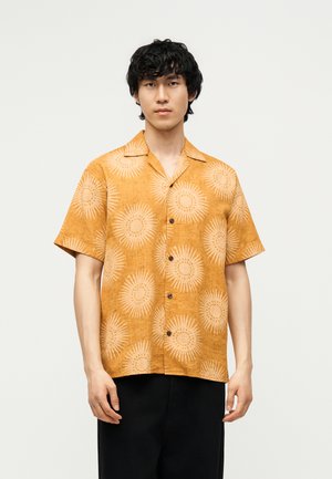 Joven con el pelo negro y rizado, vestido con una camisa naranja de manga corta con estampados de rayos de sol y pantalones negros sobre un fondo liso.