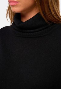Pull à col roulé noir avec un col côtelé, une texture douce et une coupe ajustée, présenté au niveau du cou et de la partie supérieure de la poitrine.