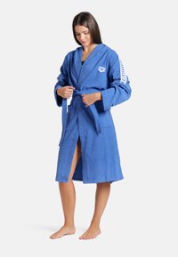 Blauwe badjas van badstof met een capuchon, lange mouwen en een strikceintuur. Bevat een geborduurd logo op de borst en een patroon op de mouwen.