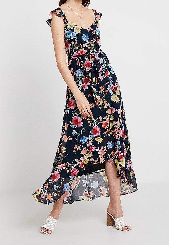 Robe maxi en bleu marine foncé avec des motifs floraux colorés, épaules à volants et ceinture à nouer. Tissu fluide avec une coupe asymétrique. Sandales à talons blanches.