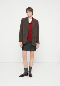 Tânără îmbrăcată cu un blazer maroniu, un cardigan roșu cu nasturi, o fustă gri închis, șosete tricotate gri și încălțăminte bordo, stând în fața unui fundal alb.