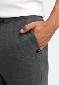 Mano masculina descansando en el bolsillo de unos pantalones gris oscuro texturizados, vistiendo una camisa blanca, con una etiqueta de tela visible cerca de la costura del bolsillo.
