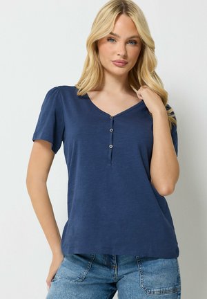 Mujer rubia con camiseta de manga corta azul marino con botones y vaqueros azul claro sobre un fondo claro y liso.