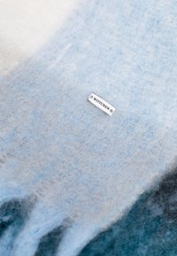 Jasnoniebieski szal z gradientem, wykonany z miękkiej, fakturowanej tkaniny; posiada srebrną metkę z nazwą marki "WITTCHEN".