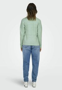 Zeleno pleteno pulover s zaobljenim robom, rebrastimi manšetami in detajli na zadnjem šivu, v kombinaciji z svetlo modrimi kavbojkami in srebrnimi čevlji.