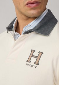 Beige pólóing sötétszürke gallérral, hímzett "H" logóval és "HACKETT" felirattal. Sima anyagból készült, hétköznapi dizájnnal.