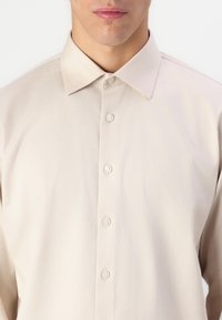 Chemise beige clair à manches longues avec un col pointu, fermeture à boutons sur le devant et texture de tissu lisse. Les boutons sont blancs et ronds.