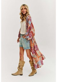 Kimono s kvetinovým vzorom v červenej, modrej a oranžovej farbe, vrstvený nad bielym pleteným tričkom, skombinovaný so svetlými džínsovými šortkami a béžovými voľnými čižmami.