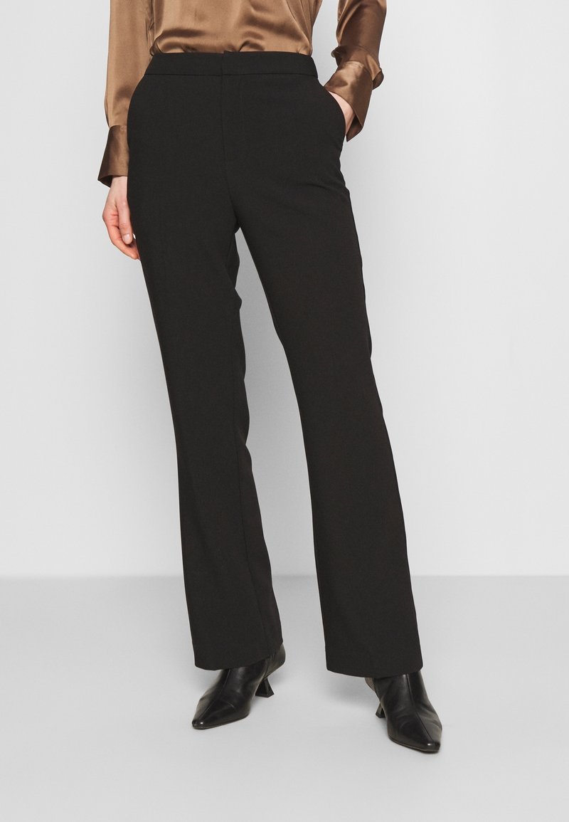 Femme portant un pantalon noir à jambes droites, un chemisier en satin marron et des bottines noires à bout pointu, debout les mains dans les poches.