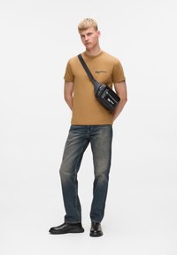 Mladý muž stojící s rukama za zády, oblečený v hořčicovém tričku, vybledlých džínách, černých botách a černé crossbody tašce na bílém pozadí.