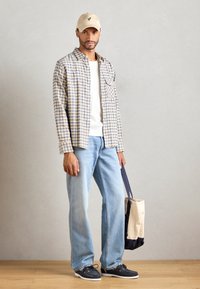 Beige caps, rutete skjorte i gult og blått, hvit t-skjorte, lyseblå jeans og mørke sko, holder en to-farget tote bag.