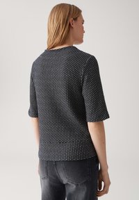 Femme aux cheveux blonds raides portant un haut à motifs noir et blanc à manches courtes et un jean gris foncé, se tenant devant un fond uni.