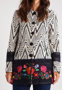 Virkat jacka med ett svart och vitt geometriskt mönster, med en blommig broderad fåll, svarta knappar och långa ärmar.