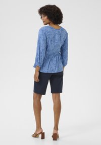 Világoskék, virágmintás blúz háromnegyedes ujjal, megkötött derékkal, sötétkék sorttal és barna blokk sarkú szandállal párosítva.