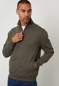 Sudadera verde oliva con cuello con cremallera y bolsillos laterales. Presenta una textura suave y puños acanalados en las muñecas y en el dobladillo.