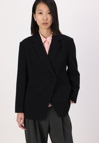 Svart blazer med strukturerte skuldre, enkelt knapplukking og to frontlommer, båret over en lys rosa skjorte og grå bukser.