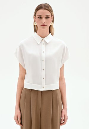 Donna che indossa una camicetta bianca a maniche corte con bottoni e colletto e pantaloni marroni a pieghe a vita alta su uno sfondo semplice.